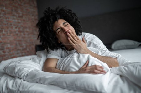 Early morning. Curly-haired young man lying in bed and yawning