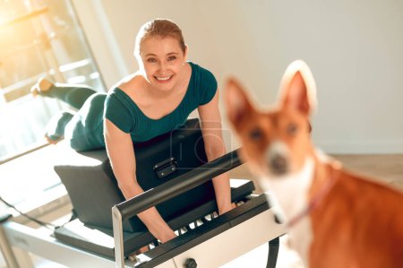 Foto de Mujer rubia sonriente haciendo ejercicios deportivos en el estudio de Pilates con su perro - Imagen libre de derechos