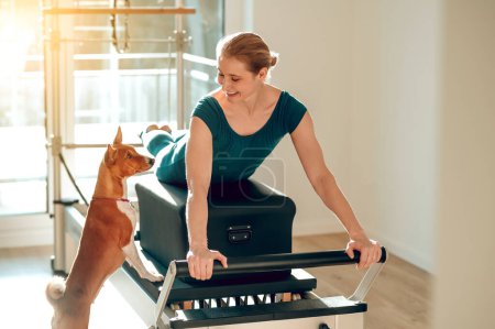 Foto de Mujer deportiva entrenando en el simulador de Pilates con su perro en el gimnasio - Imagen libre de derechos