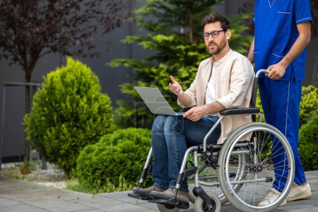 Téléchargez les photos : Connecté. Jeune homme en fauteuil roulant avec ordinateur portable dans une cour d'hôpital - en image libre de droit