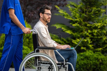 Photo for On a walk. Male nurse in blue uniform carrying a wheelchair with male patient - Royalty Free Image