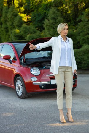 Téléchargez les photos : Femme blonde debout près de leur voiture cassée et l'autostop ayant des problèmes avec son auto sur la route - en image libre de droit