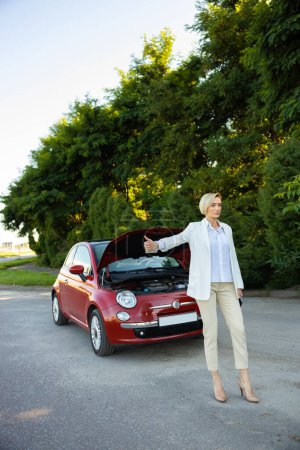 Photo for Businesswoman raises her finger while standing on country road needs help with her broken car stops drivers for help - Royalty Free Image