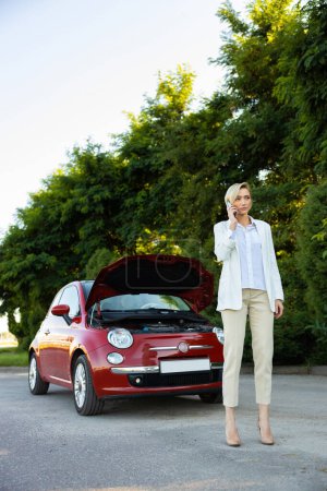 Photo for Adult businesswoman standing near broken automobile with open hood calling to service standing on road - Royalty Free Image