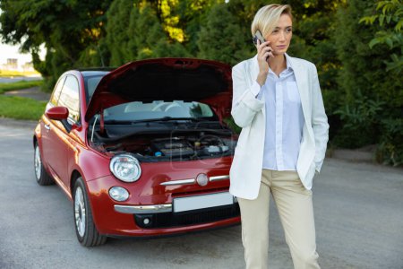 Téléchargez les photos : Femme blonde conductrice utilisant un smartphone ayant un problème avec sa voiture cassée tout en se tenant sur la route - en image libre de droit