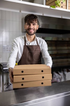 Téléchargez les photos : Homme souriant dans un tablier tenant des boîtes à pizza travaille dans la cuisine du restaurant préparant de savoureux repas italiens pour la livraison et à emporter - en image libre de droit