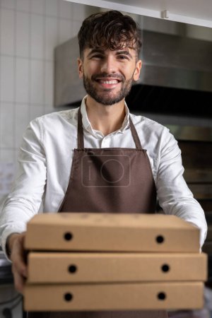 Photo for Bearded man in apron holding pizza boxes manages kitchen operations ensuring fast food service and efficient meal preparation - Royalty Free Image