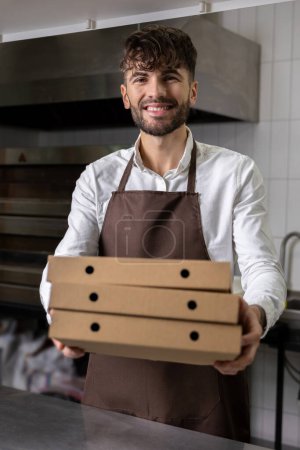 Téléchargez les photos : Satisfait homme barbu dans tablier tenant des boîtes de pizza est travailleur dans un café de pizza offrant des options de livraison express et à emporter - en image libre de droit