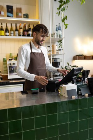 Téléchargez les photos : Serveur homme dans tablier travaillant dans un café en utilisant le terminal tout en se tenant au comptoir - en image libre de droit