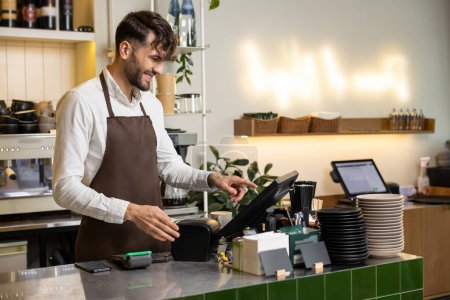 Foto de Hombre camarero en delantal trabajando en cafetería usando terminal mientras está parado en el mostrador - Imagen libre de derechos