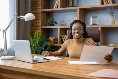 Photo for Delighted woman with dreadlocks performs remote work using laptop in home office interior - Royalty Free Image