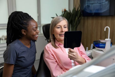 Téléchargez les photos : Patiente d'âge moyen regardant ses nouveaux implants dentaires dans un miroir à l'intérieur d'une clinique de stomatologie - en image libre de droit