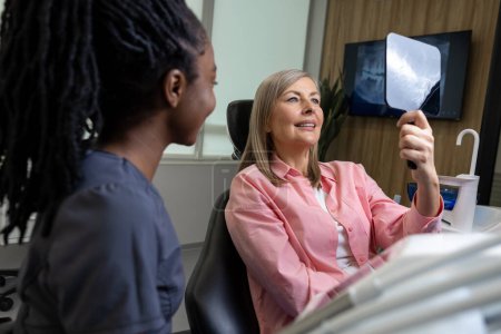 Téléchargez les photos : Patiente d'âge moyen regardant ses nouveaux implants dentaires dans un miroir à l'intérieur d'une clinique de stomatologie - en image libre de droit