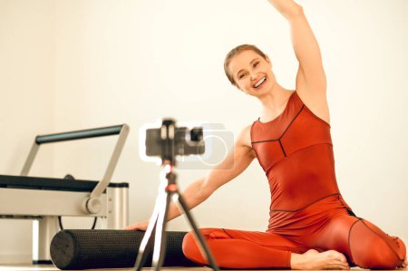 Téléchargez les photos : Magnifique instructeur de yoga femme enregistrant une formation en ligne avec smartphone et trépied dans un studio de fitness - en image libre de droit