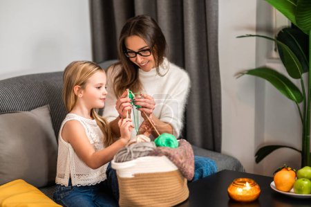 Photo for Young mother teaching her daughter to knot on sofa in home interior knitting with her daughter - Royalty Free Image