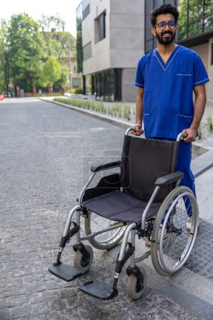 Téléchargez les photos : Homme en fauteuil roulant. Infirmière masculine positive dans les médecins bleus portant globalement un fauteuil roulant - en image libre de droit