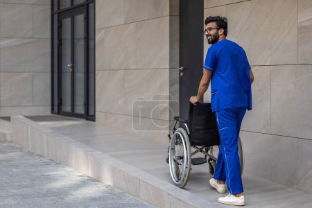 Foto de El hombre de azul médico en general. Hombre alto de cabello oscuro en el patio de los hospitales llevando una silla de ruedas - Imagen libre de derechos