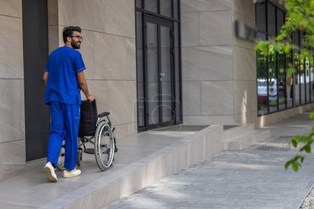Photo for Man in blue medical overall. Tall dark-haired man in hospitals yard carrying a wheelchair - Royalty Free Image