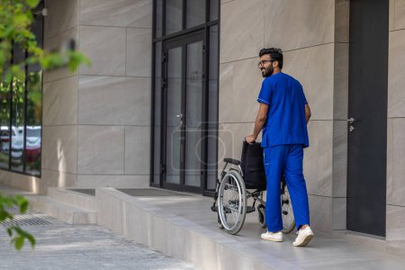 Téléchargez les photos : Homme en bleu médical en général. Grand homme brun dans la cour des hôpitaux portant un fauteuil roulant - en image libre de droit