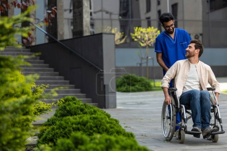 Foto de Buen humor. Enfermera masculina rodando una silla de ruedas con paciente y ambos sintiéndose bien - Imagen libre de derechos