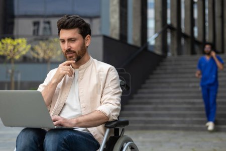 Foto de Hombre con portátil. Hombre autónomo discapacitado que trabaja en la computadora portátil mientras está sentado en silla de ruedas en el patio del hospital - Imagen libre de derechos