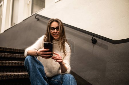 Téléchargez les photos : Jeune femme en vêtements décontractés debout sur les escaliers en utilisant le téléphone mobile défilant en ligne pendant le temps libre à l'hôtel - en image libre de droit
