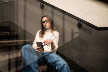 Téléchargez les photos : Attrayant femelle en utilisant le téléphone mobile pour discuter en ligne sur les escaliers de l'hôtel - en image libre de droit