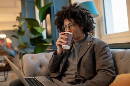 Téléchargez les photos : Café du matin. Jeune homme aux cheveux bouclés assis dans une salle d'hôtel et buvant du café - en image libre de droit