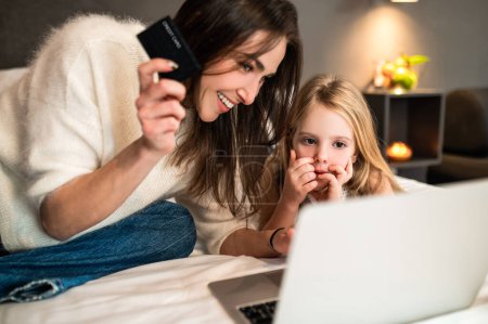 Téléchargez les photos : Femme heureuse et petite fille assise avec ordinateur portable et carte de crédit réservation en ligne de chambre d'hôtel - en image libre de droit