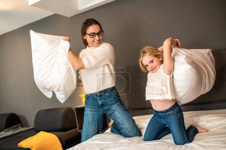 Photo for Young mother and her little daughter playing with cushions on bed in hotel room - Royalty Free Image