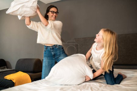 Foto de Alegre madre y su hijita disfrutando divertida pelea de almohadas jugando juntos el fin de semana - Imagen libre de derechos