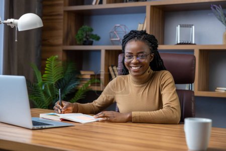 Téléchargez les photos : Femme souriante dans des lunettes avec dreadlocks assis sur le lieu de travail avec ordinateur portable dans le bureau à la maison - en image libre de droit