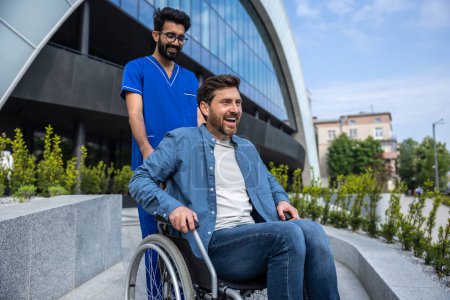 Téléchargez les photos : Bonne humeur. Jeune homme barbu souriant en fauteuil roulant faisant une promenade avec une infirmière - en image libre de droit