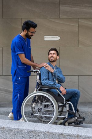 Foto de Paciente discapacitado. Enfermera morena llevando a un paciente en silla de ruedas a dar un paseo - Imagen libre de derechos