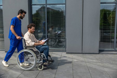 Photo for Walk with the patient. Male nurse taking a disabled patient on a wheelchair for a walk - Royalty Free Image