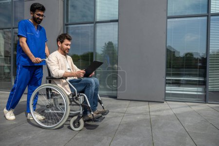 Foto de En un hospital. Hombre discapacitado dando un paseo con un enfermero y leyendo su folleto recetado - Imagen libre de derechos