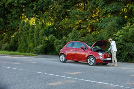 Téléchargez les photos : Femme parlant téléphone demandant de l'aide à côté de sa voiture de dépannage dans la route avec forêt verte sur fond - en image libre de droit