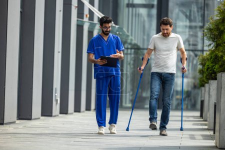 Photo for Man on rehabilitation. Young bearded man with crutches walking and talking to a male nurse - Royalty Free Image