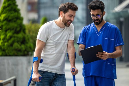 Téléchargez les photos : Réadaptation. Homme avec des béquilles et un infirmier en uniforme bleu marchant dans une cour d'hôpital - en image libre de droit