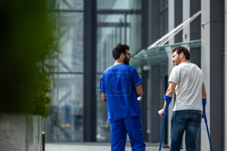 Photo for Rehabilitation. Man with crutches and a male nurse in a blue uniform walking in a hospital yard - Royalty Free Image