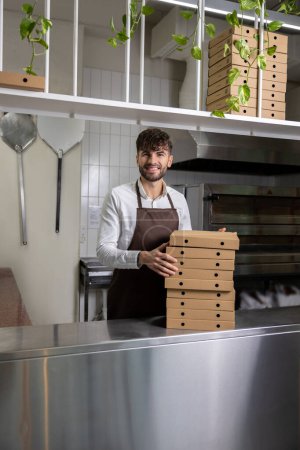Photo for Bearded man cafe employee holding stack of pizza boxes at kitchen counter - Royalty Free Image