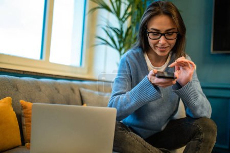 Téléchargez les photos : Femme aux cheveux bruns dans des lunettes à l'aide d'un smartphone et d'un ordinateur portable assis sur le canapé près de la fenêtre - en image libre de droit