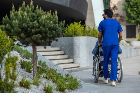 Foto de Camina con paciente. Enfermera en un médico azul llevando una silla de ruedas - Imagen libre de derechos