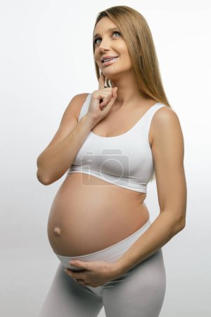 Téléchargez les photos : Une femme enceinte. Blonde femme enceinte en vêtements blancs à la recherche réfléchie - en image libre de droit