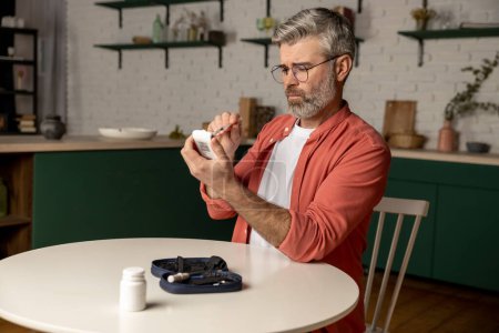 Téléchargez les photos : Homme d'âge moyen aux cheveux gris avec glucomètre vérifiant le taux de sucre dans le sang à la maison - en image libre de droit