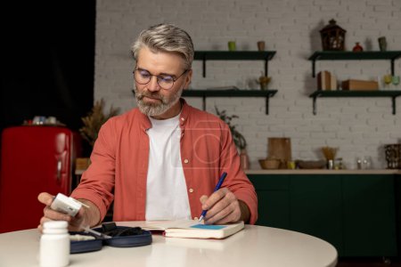 Photo for Gray haired middle aged man with glucometer checking blood sugar level taking notes with indicators in notepad at home - Royalty Free Image