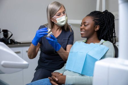 Téléchargez les photos : Dentiste féminine montrant des prothèses dentaires à sa patiente à la clinique - en image libre de droit
