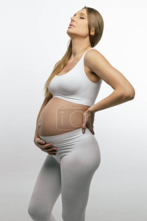 Foto de Embarazo. Mujer embarazada con la parte superior blanca y polainas sosteniendo su mano sobre su vientre - Imagen libre de derechos