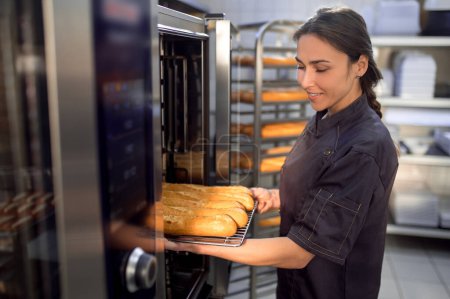 Téléchargez les photos : Femme boulangerie utilisant four électrique pour la cuisson des baguettes françaises dans la boulangerie - en image libre de droit