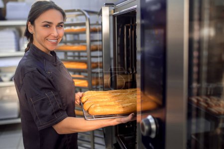 Foto de Retrato de una joven panadera en uniforme horneando baguettes frescas en la panadería - Imagen libre de derechos
